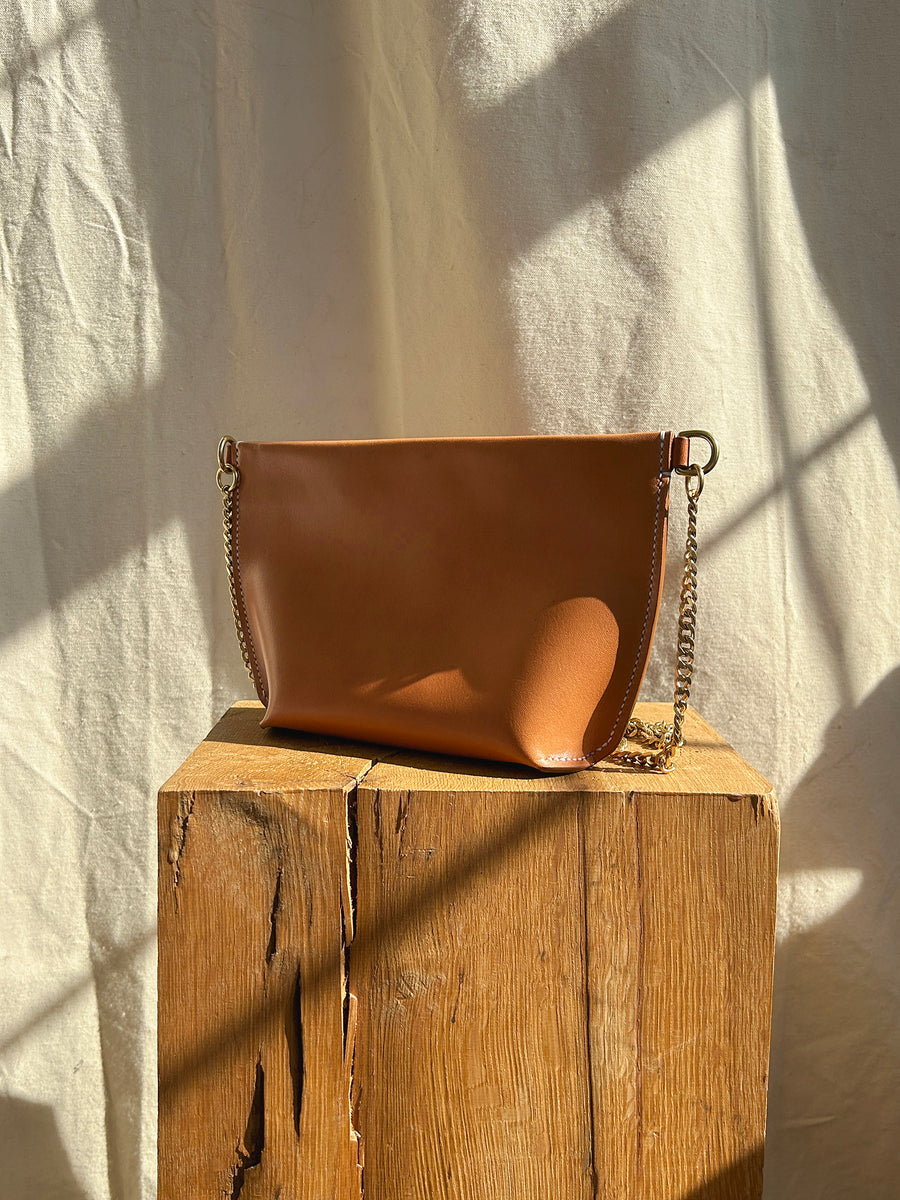 The Bartleby Objects Stowe Chain Bag, crafted from vegetable-tanned brown leather with a chain strap, sits on a wooden surface. The textured background is illuminated by sunlight, casting shadows on the bag and creating a warm, natural atmosphere. This saddle-stitched bag exudes artisanal craftsmanship.