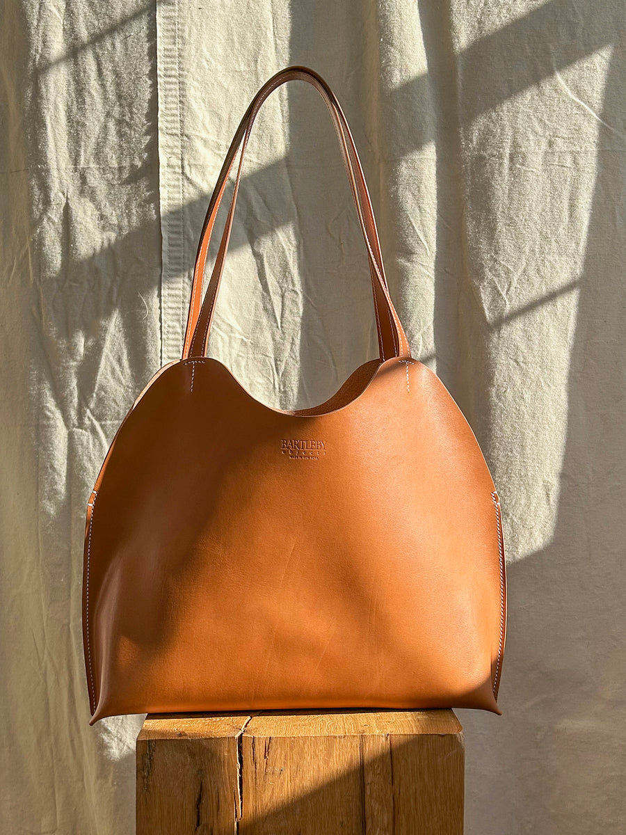 The Bartleby Objects Freja Tote, crafted from vegetable-tanned leather, features two handles and rests on a wooden block. The textured, light-colored fabric in the background casts shadows that enhance its elegant and classic ambiance. Handmade in New York City by Bartleby Objects, this tote exudes sophistication.