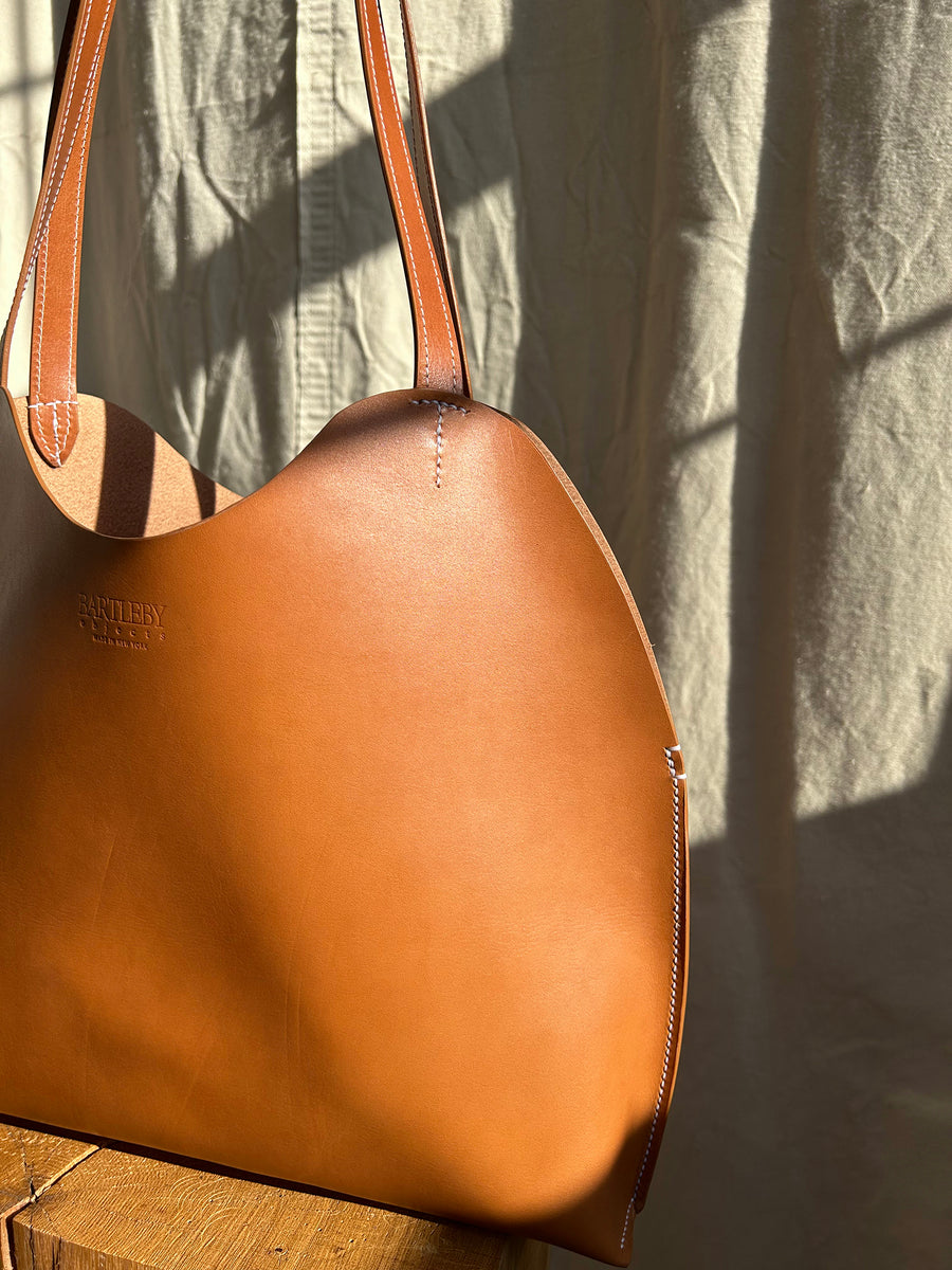 The Bartleby Objects Freja Tote, crafted from vegetable-tanned leather with brown straps, hangs elegantly on a wooden stand. Sunlight softly graces the textured beige curtain in the background, creating a warm and inviting atmosphere. This expertly handmade piece is produced in New York City by Bartleby Objects.