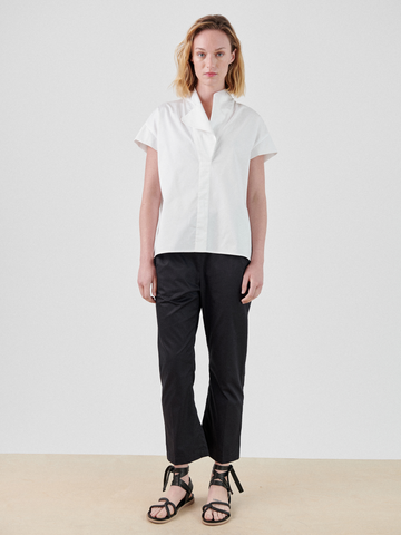 A person with shoulder-length hair stands facing the camera. They are wearing a white, crisp woven cotton blouse, black Eko Pants by Zero + Maria Cornejo, and black strappy sandals. The background is plain and neutral.