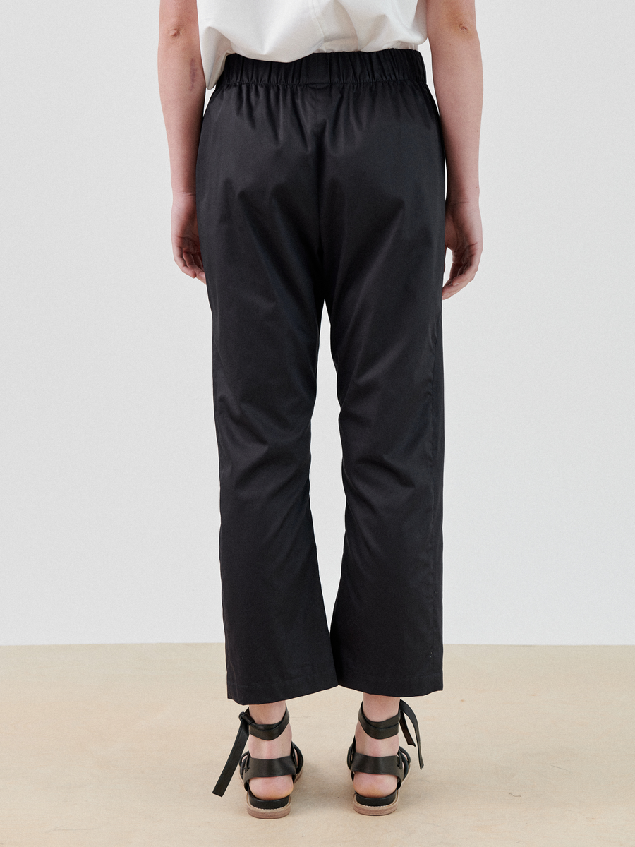 A person wearing a white shirt and black Zero + Maria Cornejo Eko Pant with an elastic waistband stands facing away from the camera. The trousers appear to be made of crisp woven cotton. They are also wearing black sandals with ankle straps. The background is plain and light-colored.