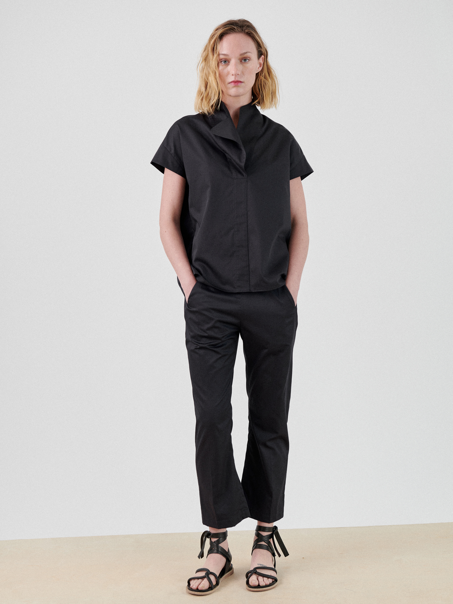 A person with shoulder-length blonde hair is standing against a plain white background. They are wearing a black short-sleeved Adi Top by Zero + Maria Cornejo made of cotton shirting and black pants, with their hands in their pockets. They are also wearing black strappy sandals.