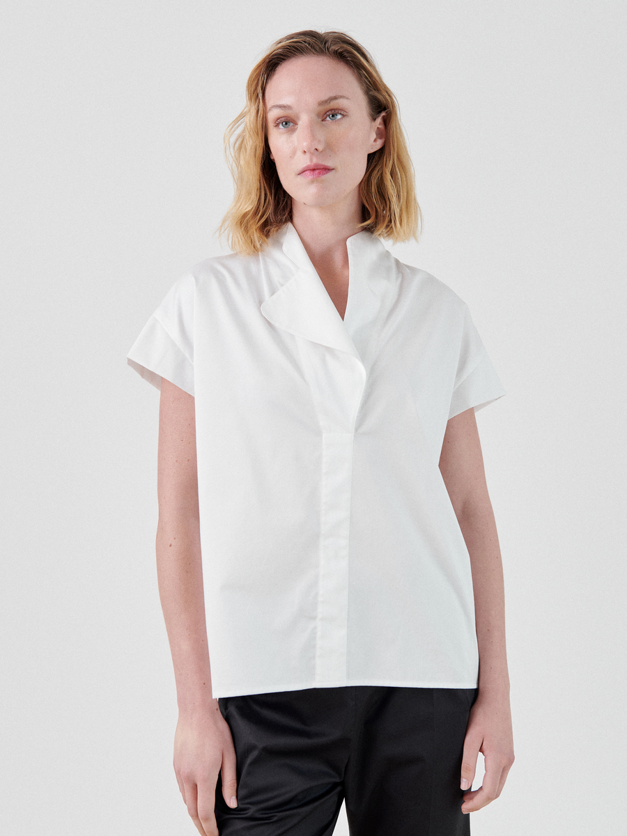 A person with shoulder-length blonde hair is wearing a white, short-sleeve, stand collar Adi Top by Zero + Maria Cornejo paired with black pants. The individual is standing against a plain white background and looking slightly off-camera with a neutral expression.