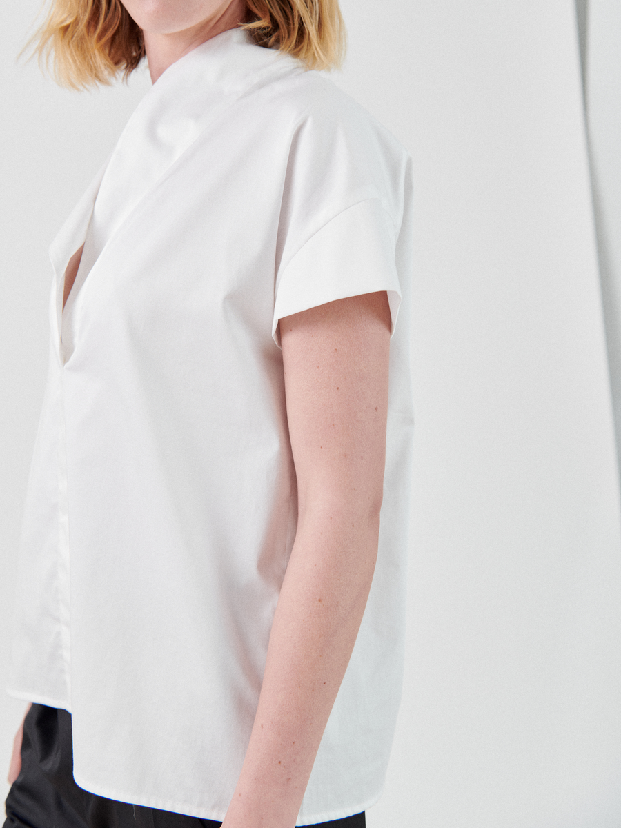 A woman in a short-sleeved, white cotton Adi Top by Zero + Maria Cornejo stands confidently, her crisp attire accentuated by a stylish stand collar.