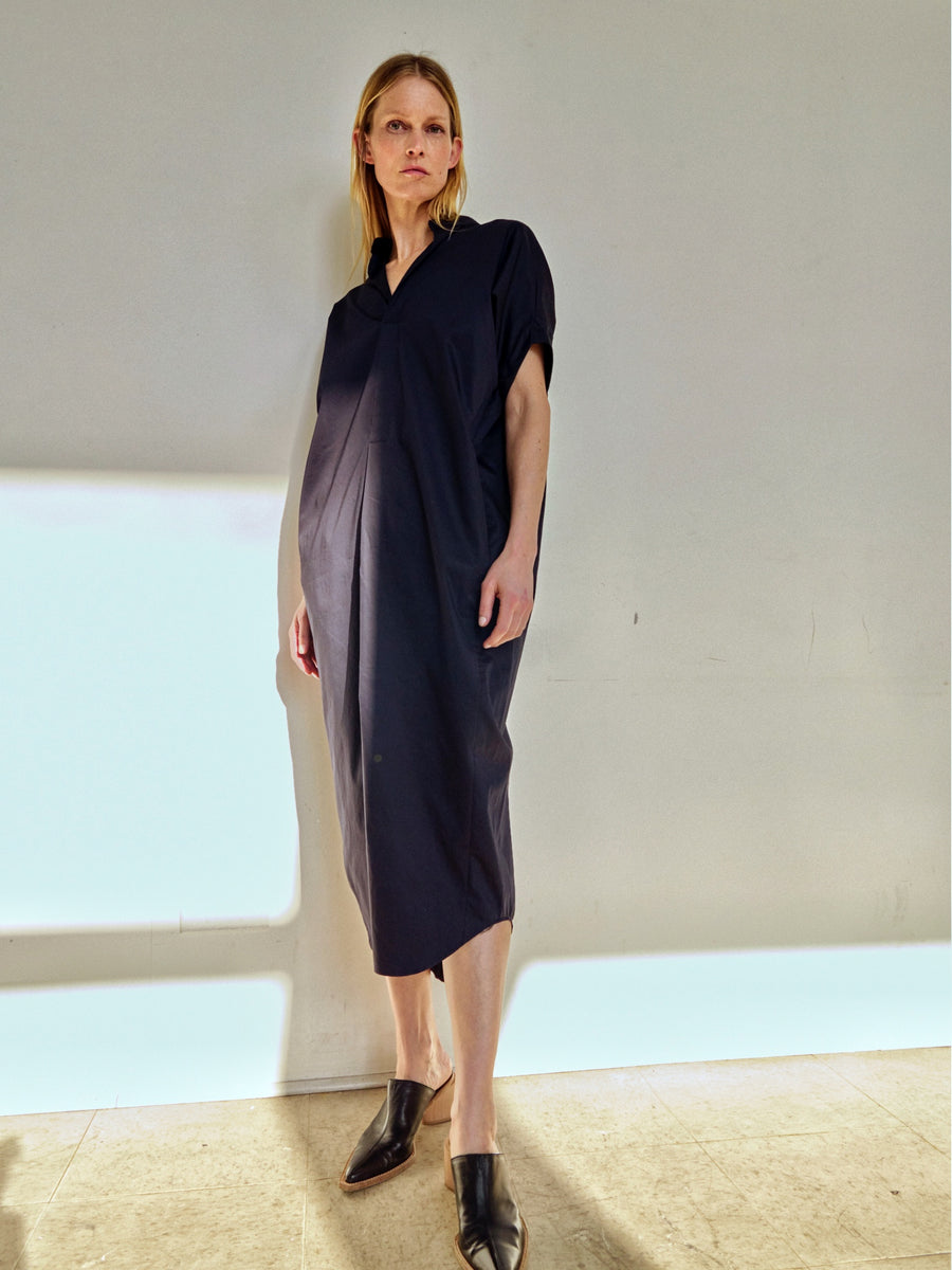 A person with shoulder-length blonde hair stands against a plain wall, wearing the Long Aissa Shirt Dress by Zero + Maria Cornejo. The loose, dark-colored dress falls below the knee, paired with black pointed-toe shoes. The draped silhouette adds elegance as they look slightly away from the camera with a neutral expression. Soft lighting casts gentle shadows around them.