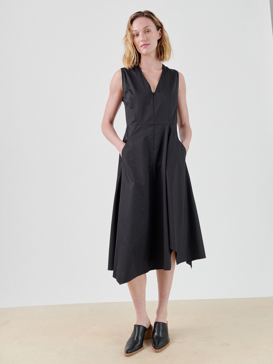 A person with shoulder-length hair stands against a plain background, wearing a Sleeveless Wave Dress by Zero + Maria Cornejo with pockets and black slip-on shoes. The mid-length dress has a flowing, slightly asymmetrical hemline. The person is looking slightly to the side.