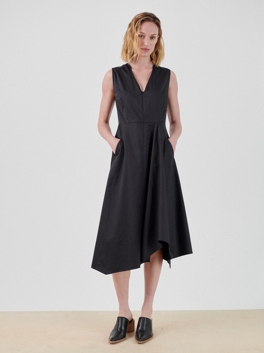 A woman stands against a plain white background, wearing the Sleeveless Wave Dress by Zero + Maria Cornejo. She has short, wavy blonde hair and is wearing black slip-on shoes. Her hands are in her dress pockets, and she gazes directly at the camera. The v-neck dress is made in New York.