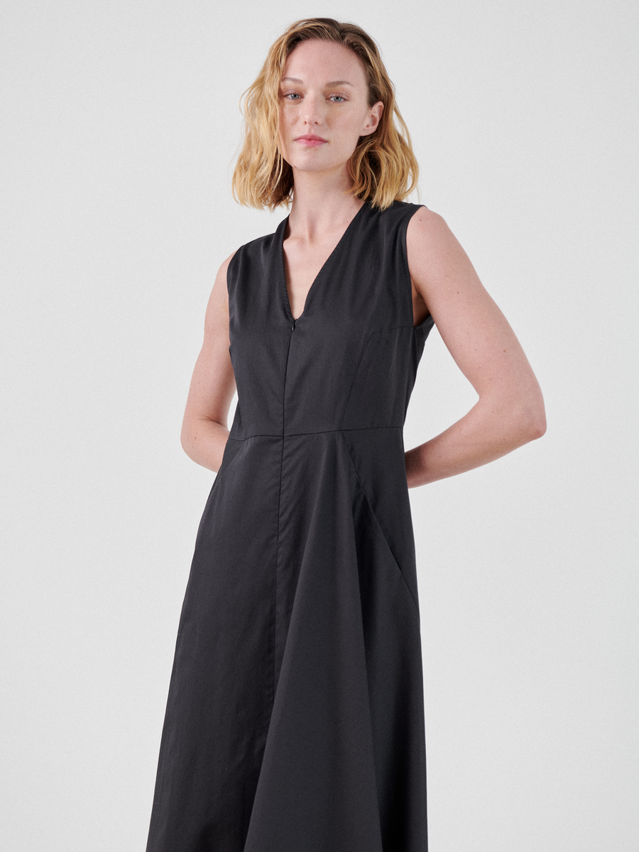 A person with shoulder-length blonde hair is wearing a Sleeveless Wave Dress by Zero + Maria Cornejo. They stand against a plain white background, looking directly at the camera with their hands behind their back. The v-neck dress has a flowy, mid-length skirt.