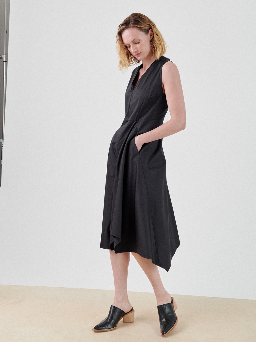 A person with shoulder-length hair wears a Sleeveless Wave Dress by Zero + Maria Cornejo. They have one hand in a pocket and are looking down. They are wearing black open-toe mules with a low heel. The background is plain and light-colored.