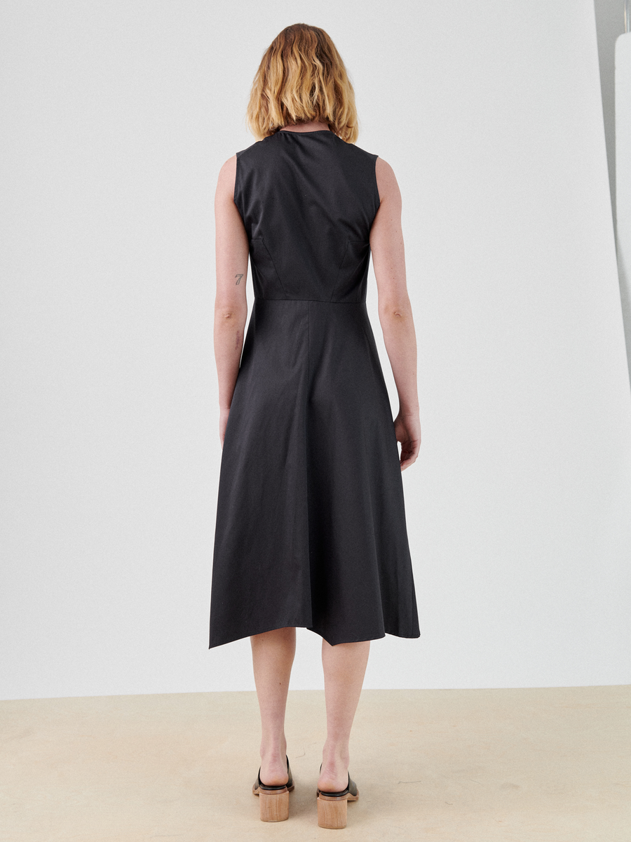 A person with shoulder-length hair is shown from behind, wearing a sleeveless, black Sleeveless Wave Dress by Zero + Maria Cornejo. They are standing on a beige surface against a plain white background. The individual is also wearing brown, open-toe sandals with a heel.