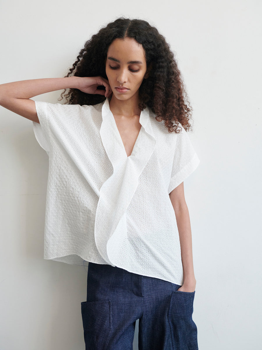 A person with long curly hair is wearing a Fin Gaban Shirt from Zero + Maria Cornejo, paired with dark pants. Their right hand is touching their neck, and their left hand is in their pants pocket. The background is a plain white wall.