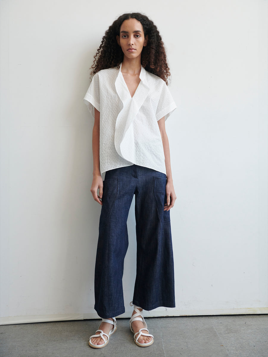 A woman with curly hair stands against a plain backdrop, wearing a white, short-sleeve, textured Fin Gaban Shirt by Zero + Maria Cornejo and loose-fitting dark blue jeans. This perfect example of summer shirting is completed with white strappy sandals. She faces the camera with a neutral expression.