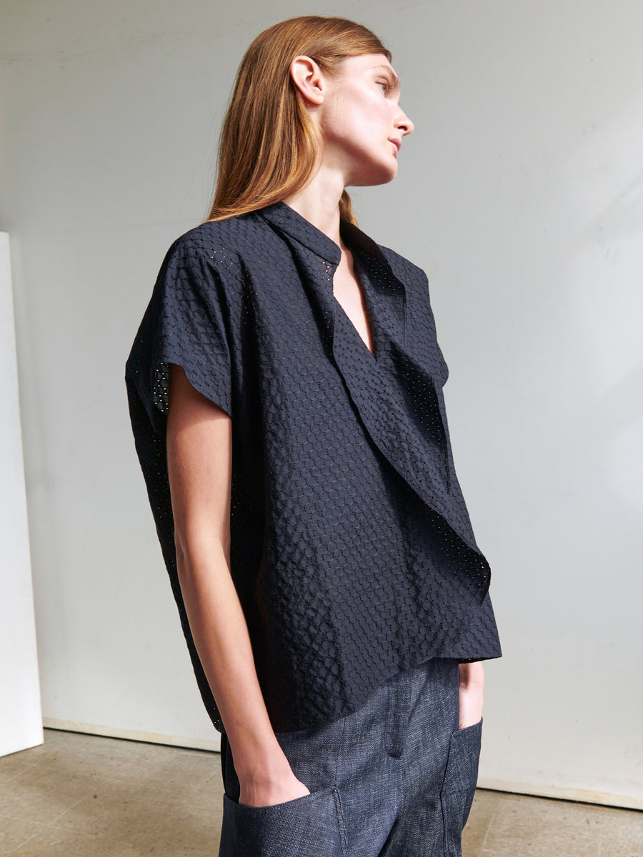 A woman with long brown hair is standing sideways, looking slightly upwards. She is wearing the Fin Gaban Shirt from Zero + Maria Cornejo, which features a textured, loose-fitting embroidered cotton design with a V-neck and short sleeves. She has paired it with dark pants. The background is a simple, neutral-toned indoor setting.