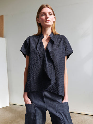 A woman with long, straight hair stands with her hands in the pockets of her denim pants. She is wearing the Fin Gaban Shirt by Zero + Maria Cornejo, a short-sleeved, textured black blouse featuring a subtle draped ruffle detail. The background is minimalistic, adorned with white and neutral tones.