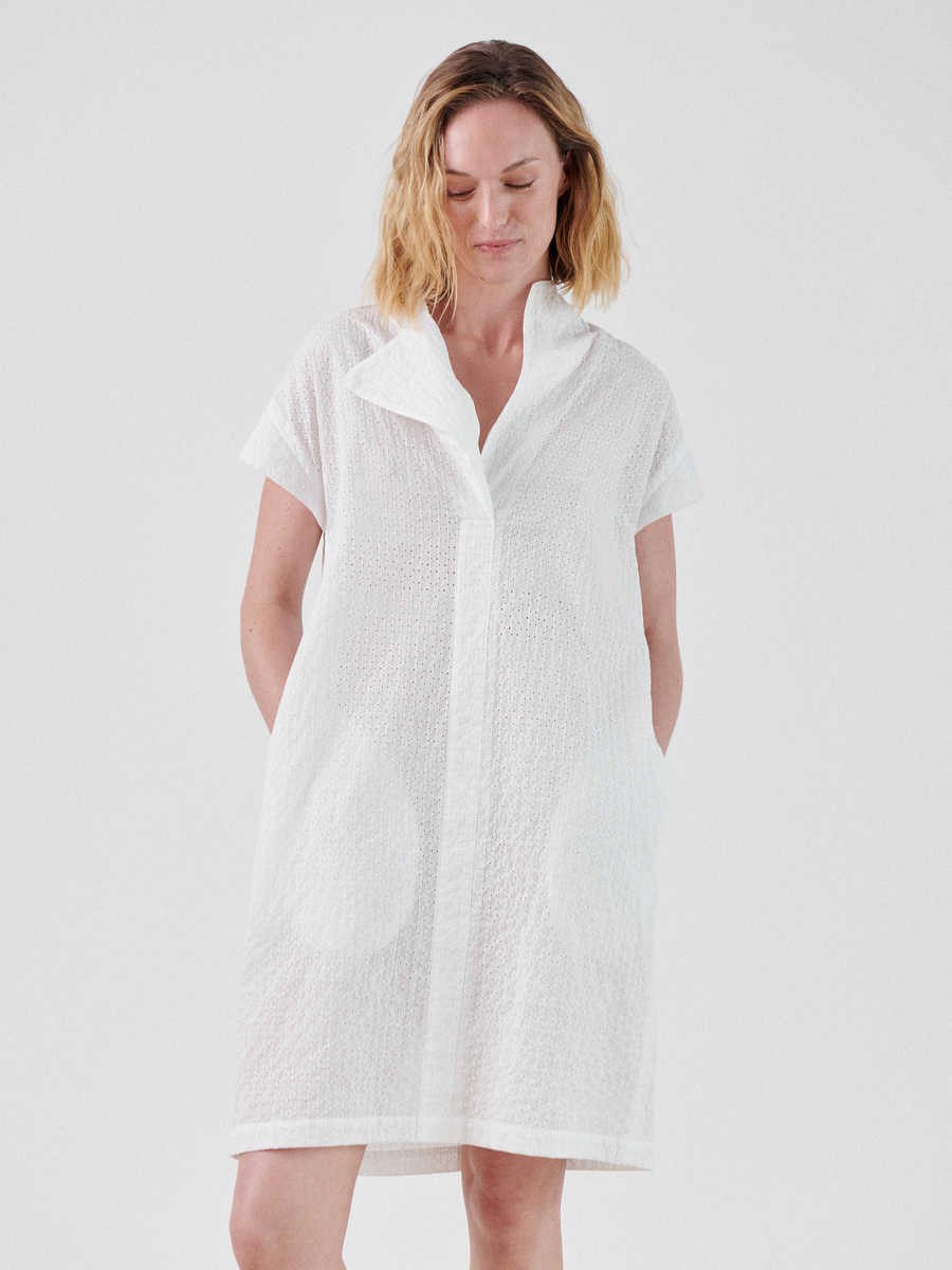 A person with shoulder-length blonde hair stands against a plain background, wearing a loose, short-sleeved white cotton Adi Dress with textured fabric by Zero + Maria Cornejo. The dress has a collar and two pockets. The person is looking slightly downward with hands in their pockets, evoking a relaxed summer shirting vibe.