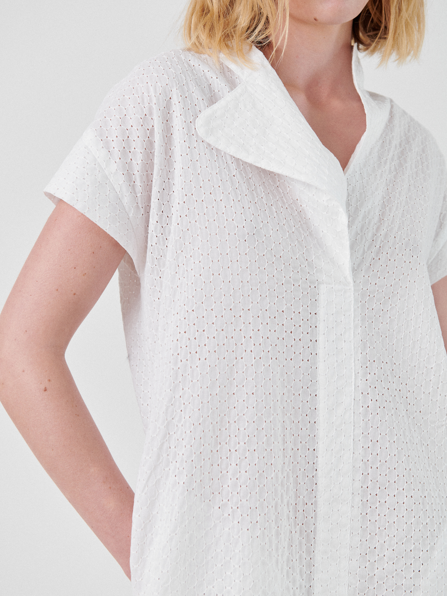 A person wearing the Adi Dress, from Zero + Maria Cornejo, with a unique asymmetrical collar, reminiscent of summer shirting. The dress features a perforated, diamond-patterned fabric. Their hand is placed in their pocket, and only part of their face is visible.