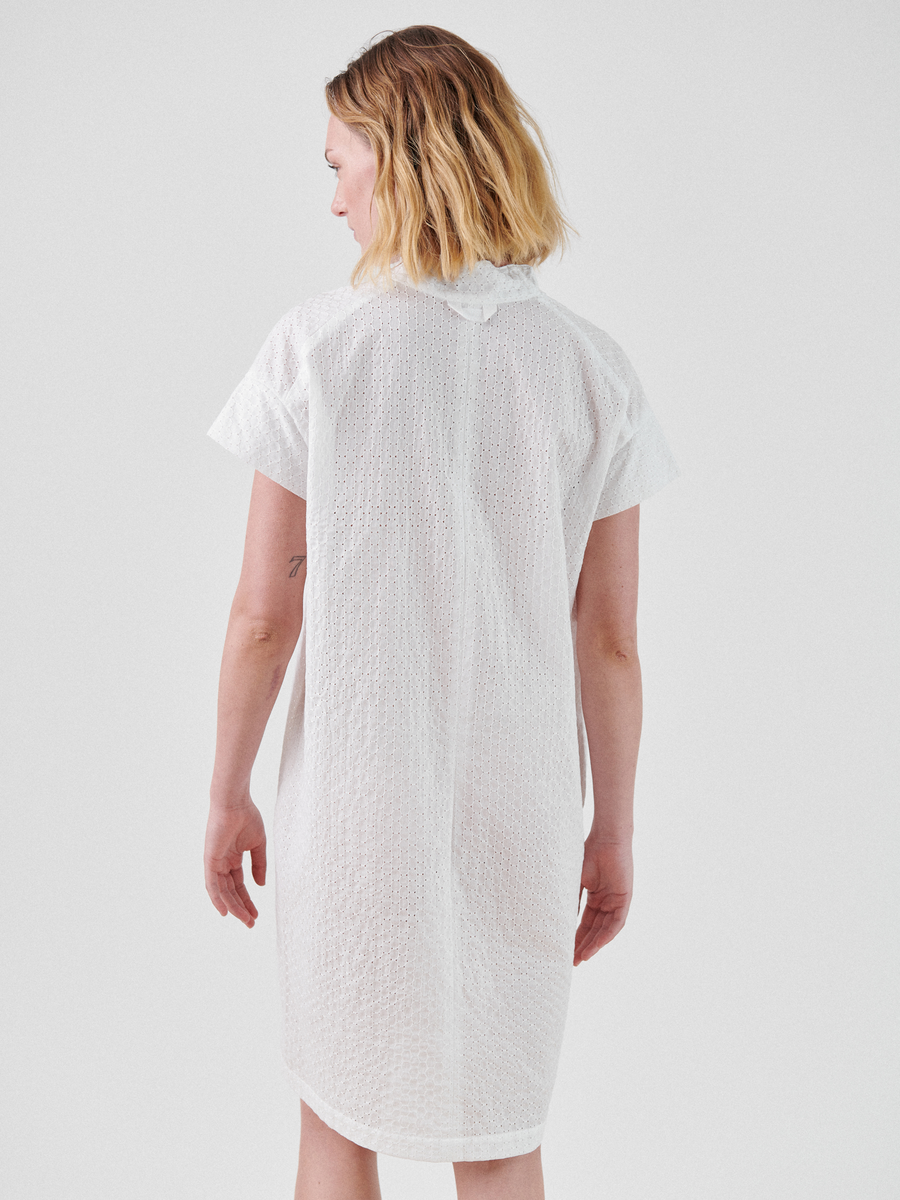 A person with shoulder-length blonde hair is standing with their back to the camera, wearing the Adi Dress by Zero + Maria Cornejo. The lightweight, textured white fabric of the summer shirting enhances their profile against the plain white background.