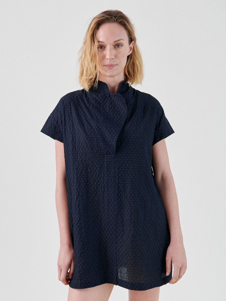 A person with shoulder-length blonde hair is standing against a plain white background. They are wearing the Adi Dress by Zero + Maria Cornejo. The textured navy blue fabric complements their relaxed expression as their right hand rests by their side.