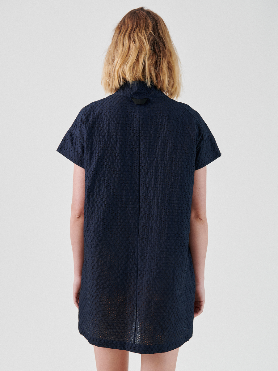 A person with blonde hair stands facing away from the camera, wearing a short-sleeved, dark blue textured cotton Adi Dress by Zero + Maria Cornejo. The dress has a loose fit and extends to just above the knees. The background is plain and light-colored, highlighting the casual summer shirting style.