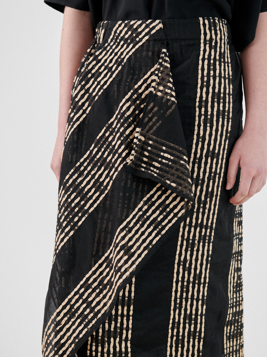 A person is wearing the Zero + Maria Cornejo One Skirt with vertical and diagonal beige stripe patterns. This summer staple features a unique draped design and asymmetrical ruffle, creating layered effects. Only the waist to mid-thigh of the person is visible against a plain, light-colored background.