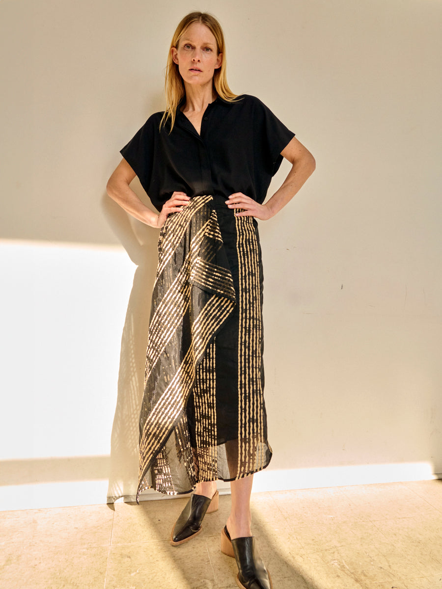 A person with long blonde hair stands against a plain background. They are wearing a black, short-sleeved blouse and an organic cotton Zero + Maria Cornejo One Skirt featuring gold, vertical and diagonal stripes. With one hand on their hip, they are looking directly at the camera—an effortless summer staple.