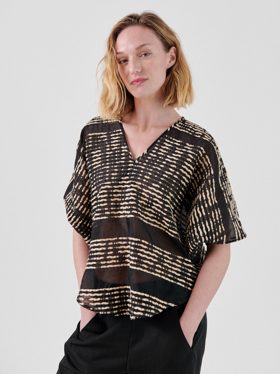 A person with shoulder-length blonde hair wears a loose-fitting, short-sleeved black Circle Top with an abstract beige pattern by Zero + Maria Cornejo. This lightweight top, made from organic cotton, is perfect as a summer staple. They are standing with their hands in their pockets against a plain white background.