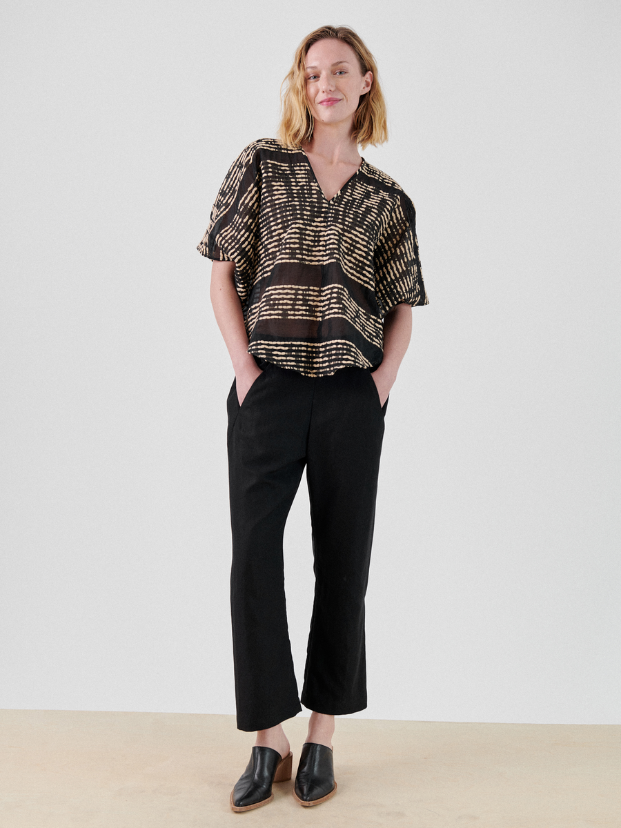 A woman with shoulder-length blonde hair is wearing a loose, short-sleeved black and beige-patterned Circle Top from Zero + Maria Cornejo, made from organic cotton, paired with high-waisted, cropped black pants. She stands casually with her hands in her pockets and is wearing black slip-on shoes. The background is plain white.