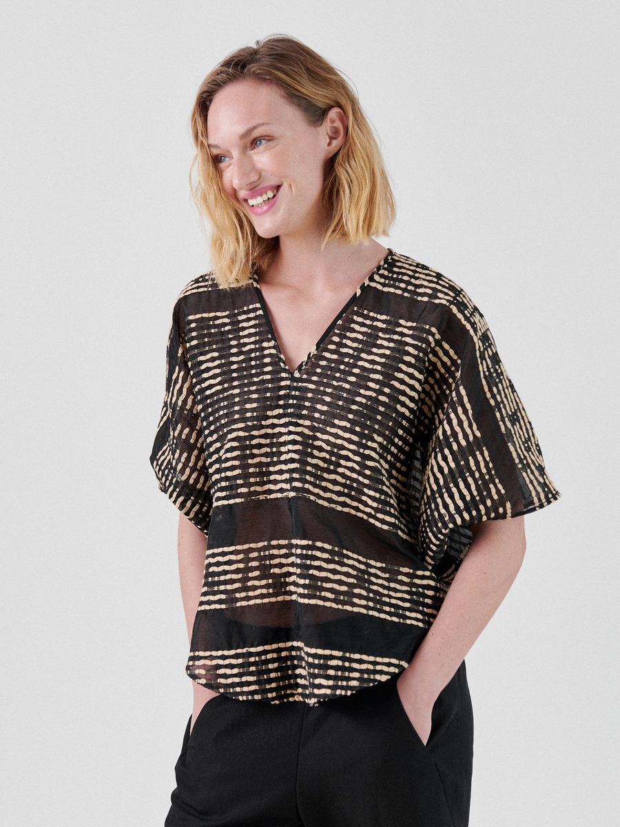 A person with shoulder-length blond hair smiles while looking off camera. They wear a black and beige patterned organic cotton Circle Top by Zero + Maria Cornejo and black pants, standing against a plain white background with hands in their pockets.