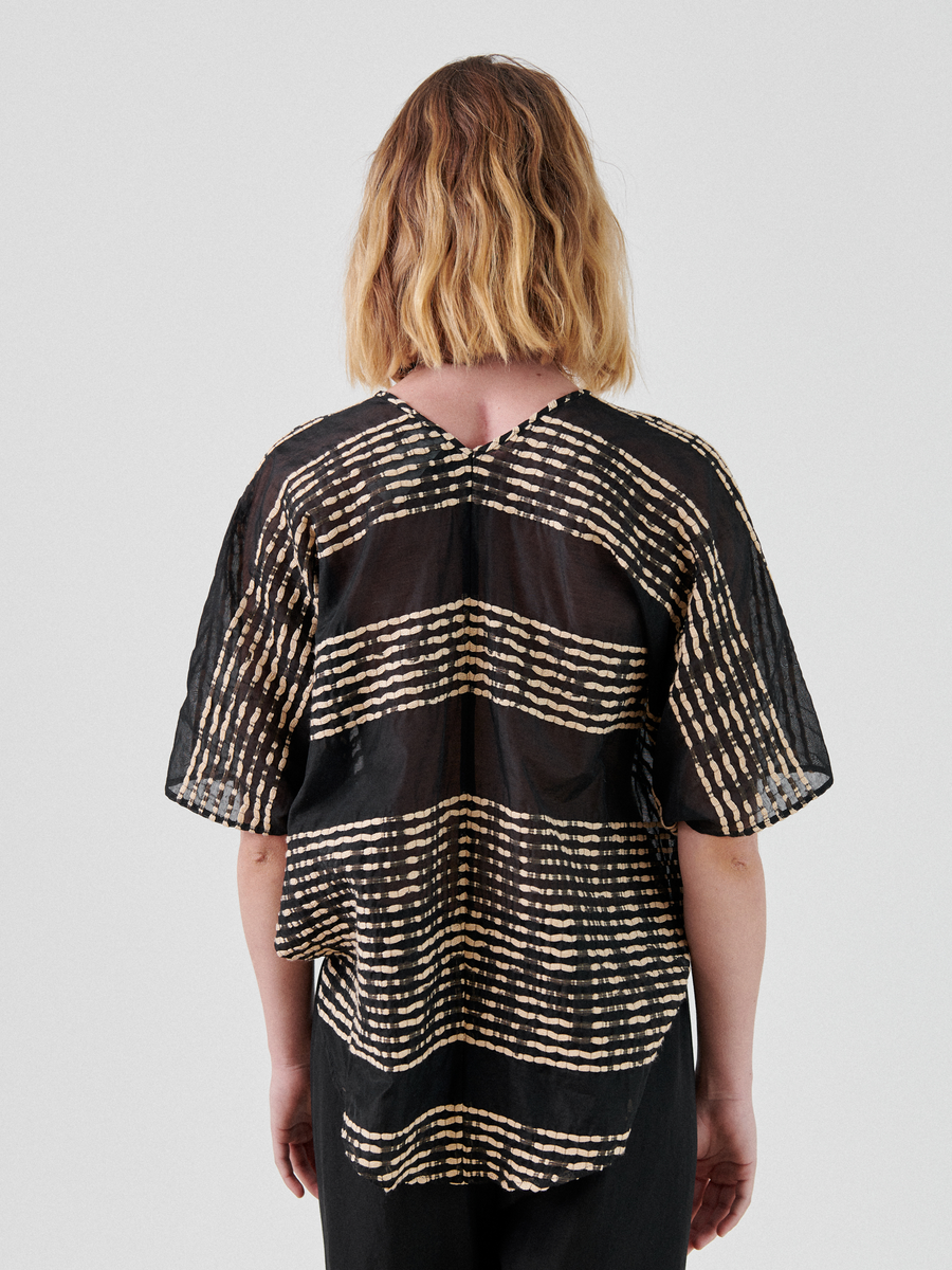 A person with shoulder-length blonde hair is seen from the back, wearing a black Zero + Maria Cornejo Circle Top with beige horizontal stripe patterns. The lightweight top has a loose fit and sheer fabric, paired with dark pants. This summer staple stands out against the plain light gray background.