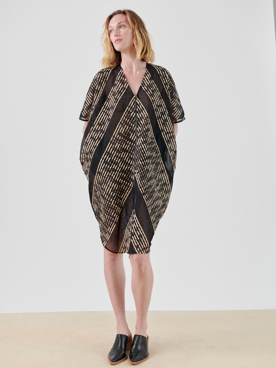 A person with shoulder-length hair stands against a neutral background wearing the Zero + Maria Cornejo Zip Caftan in an abstract black and yellow striped pattern. This summer staple, crafted from organic cotton, features handy pockets and is paired with black slip-on shoes.