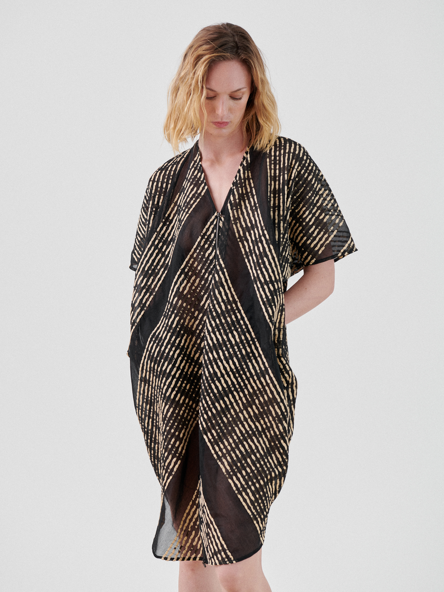 A woman with shoulder-length blonde hair wears a loose, short-sleeved Zip Caftan by Zero + Maria Cornejo featuring a black and tan diagonal striped pattern. This chic summer staple is made from organic cotton. She looks down with her hands in her pockets, standing against a plain white background.