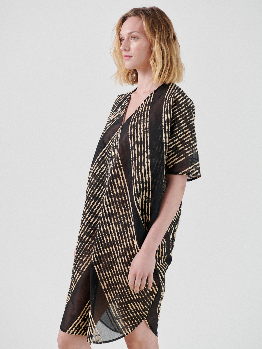 A person with shoulder-length blonde hair wears a knee-length, loose-fitting woven Zip Caftan from Zero + Maria Cornejo made from organic cotton, featuring a geometric black and tan pattern. The lightweight caftan has short sleeves and a V-neckline. The individual stands against a plain, light grey background.