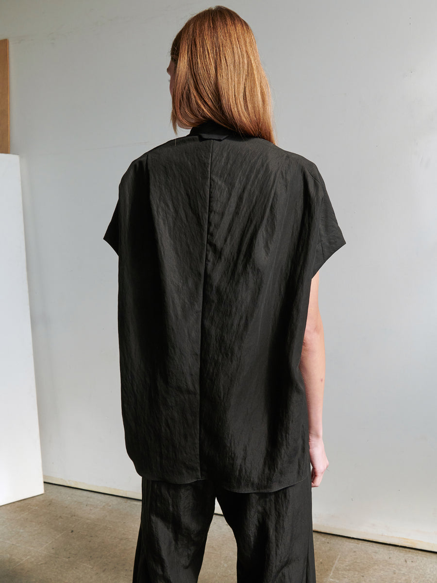 A person with long, light brown hair stands facing away from the camera, wearing the loose-fitting black Fin Gaban Shirt with short sleeves and matching black pants from Zero + Maria Cornejo. The technical woven shirt features a stand collar and is made from sustainable TENCEL™ Lyocell. The background is minimalistic with neutral tones.