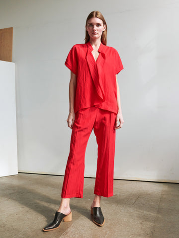 A person with long hair is dressed in a loose-fitting, vibrant red outfit from Zero + Maria Cornejo, featuring the Eko Pant with a wide banded elastic waist and a matching short-sleeve top. They stand against a plain, light-colored background, wearing black mule-style shoes. The floor beneath them is polished concrete.