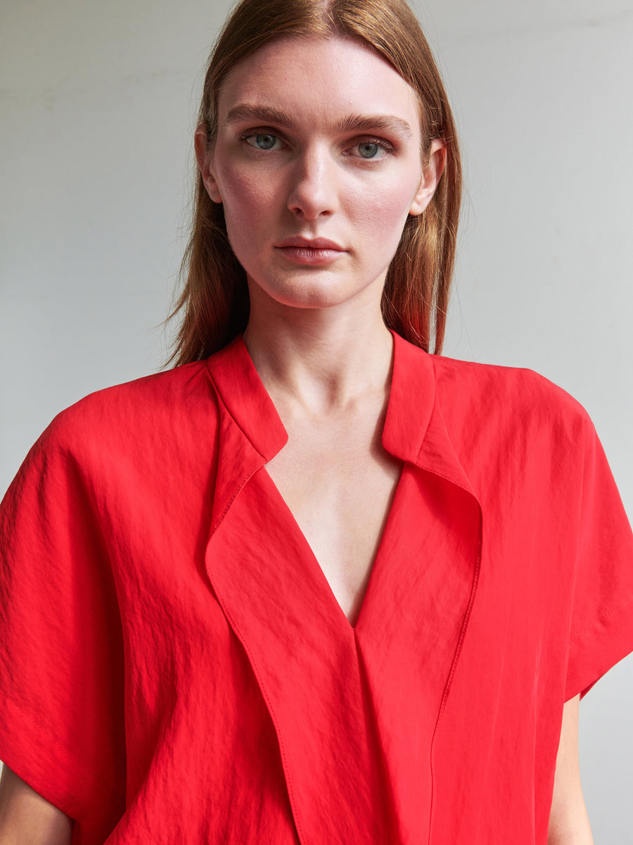 A person with straight, brown hair and light skin wearing a bright red Fin Gaban Shirt from Zero + Maria Cornejo, featuring a v-neck with draped ruffle detail, gazes directly into the camera with a neutral expression. The background is plain and unfocused, putting emphasis on the individual's face and upper body.