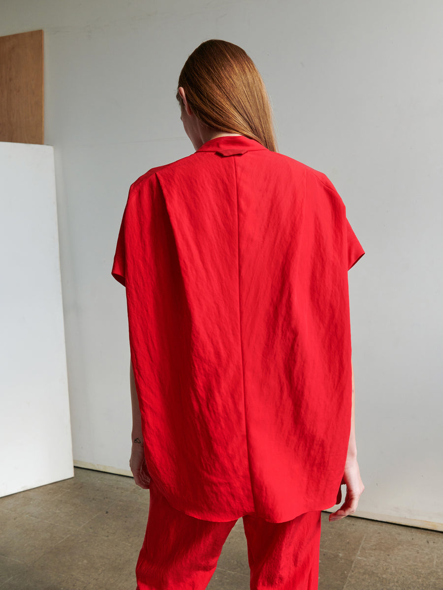 A person with long, brown hair stands facing away from the camera, wearing a loose Fin Gaban Shirt and matching red pants by Zero + Maria Cornejo. The background is a minimalist room with neutral-colored walls and a concrete floor.