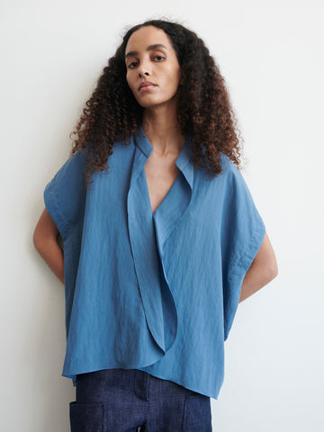 A person with long, curly hair is posing against a plain background, wearing a loose-fitting blue Fin Gaban Shirt by Zero + Maria Cornejo with short sleeves and an asymmetrical neckline that features a draped ruffle detail. Their hands are positioned behind their back.