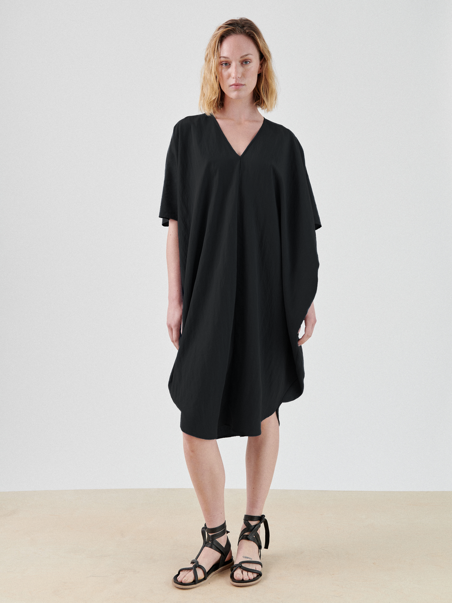 A person with shoulder-length hair is standing against a plain background, wearing a loose-fitting, knee-length Circe Dress by Zero + Maria Cornejo with a V-neckline and short, wide sleeves. They also wear black strapped sandals.