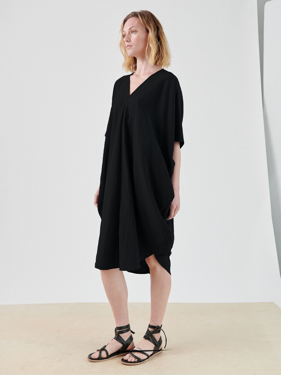 A person with shoulder-length blonde hair stands wearing a knee-length, short-sleeved black Circe Dress by Zero + Maria Cornejo made from TENCEL™ Lyocell and black gladiator sandals. They are standing on a neutral-toned surface with a plain white background, gazing slightly to the left.