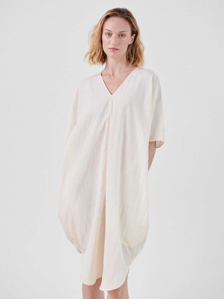 A woman with shoulder-length blonde hair stands against a plain background, wearing a loose-fitting, off-white Circe Dress made from TENCEL™ Lyocell with short sleeves by Zero + Maria Cornejo. Her hands are concealed behind the dress, and she gazes directly at the camera.