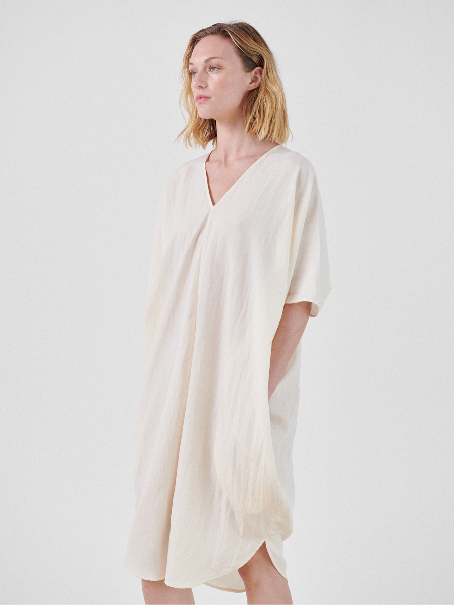 A person with shoulder-length blonde hair is wearing a loose-fitting, knee-length Circe Dress from Zero + Maria Cornejo made from TENCEL™ Lyocell with a V-neckline. They are standing against a plain, light-colored background, with one hand in a pocket and looking to the side.