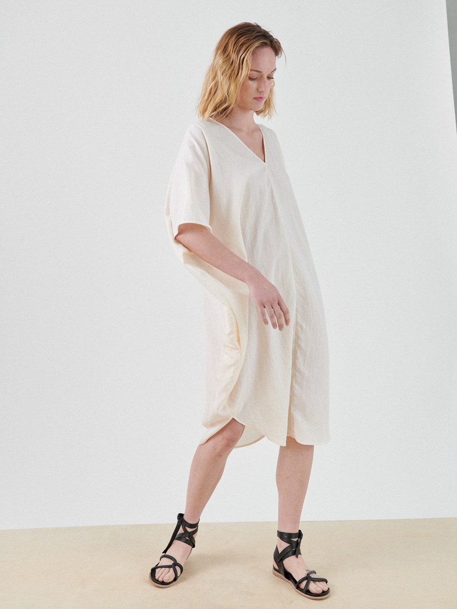 A person with shoulder-length, wavy hair is wearing a loose, light beige Circe Dress by Zero + Maria Cornejo and black strappy sandals. They are standing against a plain, light-colored background, looking down with a relaxed posture.