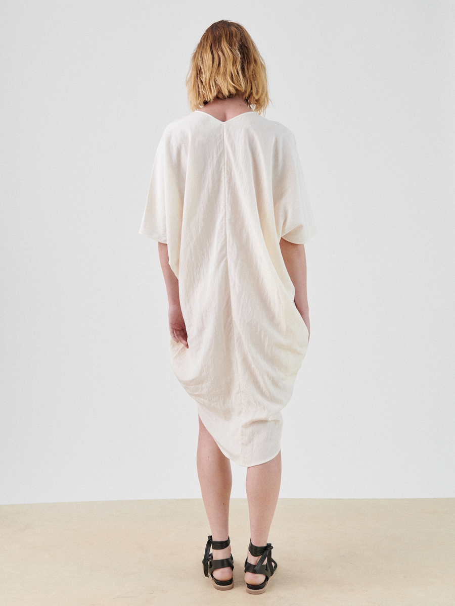 A person with shoulder-length blonde hair is standing with their back facing the camera, wearing a loose-fitting technical woven white Circe Dress by Zero + Maria Cornejo that reaches below the knees and black sandals. The background is plain and light-colored.