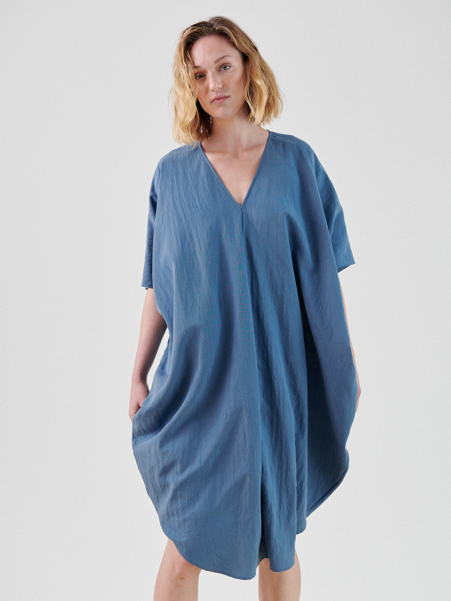 A person with blonde hair is standing against a plain white background, wearing a loose-fitting, blue Circe Dress by Zero + Maria Cornejo made of TENCEL™ Lyocell with short sleeves. The person's hands are relaxed at their sides, and they are looking directly into the camera with a neutral facial expression.