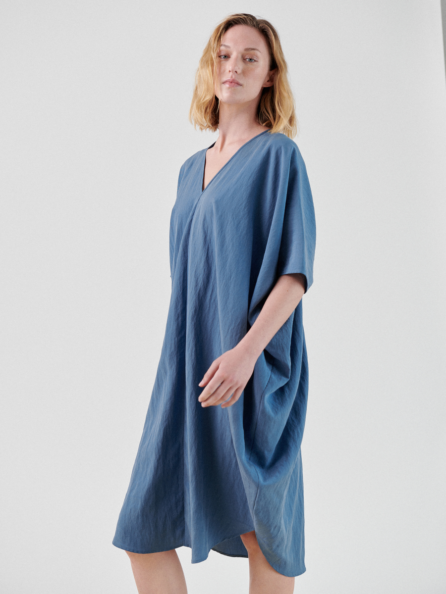 A person with shoulder-length blonde hair is wearing a loose-fitting, knee-length Circe Dress by Zero + Maria Cornejo. They are standing against a plain white background and gazing forward with a relaxed expression. The blue dress has short sleeves, a V-neck, and falls just above the knees.