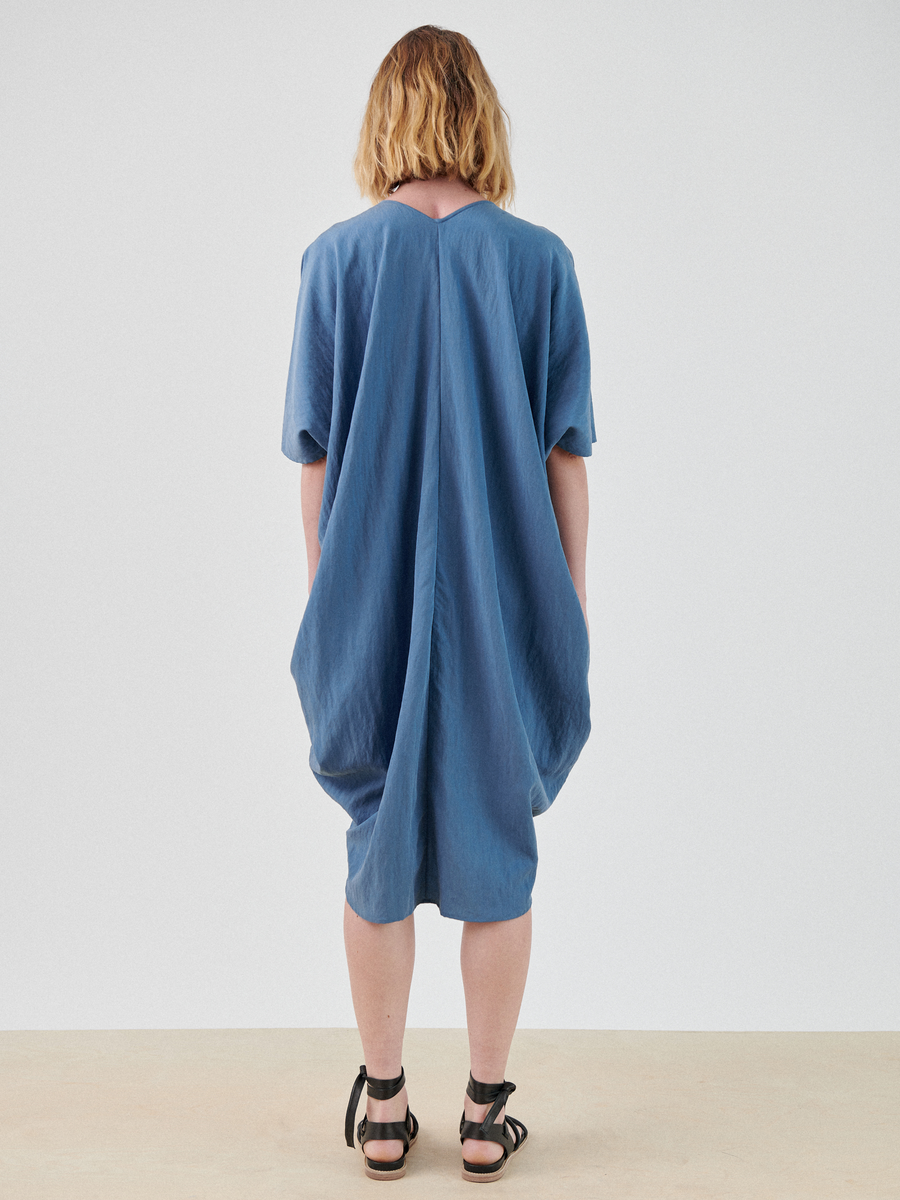 A person with shoulder-length hair stands facing away from the camera, wearing a loose, blue Circe Dress by Zero + Maria Cornejo made from TENCEL™ Lyocell and black sandals. The dress has a relaxed fit, with draped fabric at the back. The background is plain and light-colored.