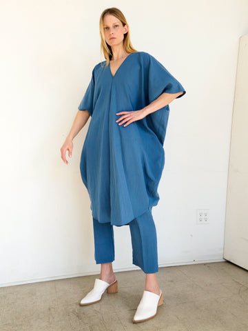 A person with long hair is standing in a minimalist room, wearing a Circe Dress from Zero + Maria Cornejo over matching blue pants and white heeled mules. The floor is plain, and the background is a simple, off-white wall with an electrical outlet visible.