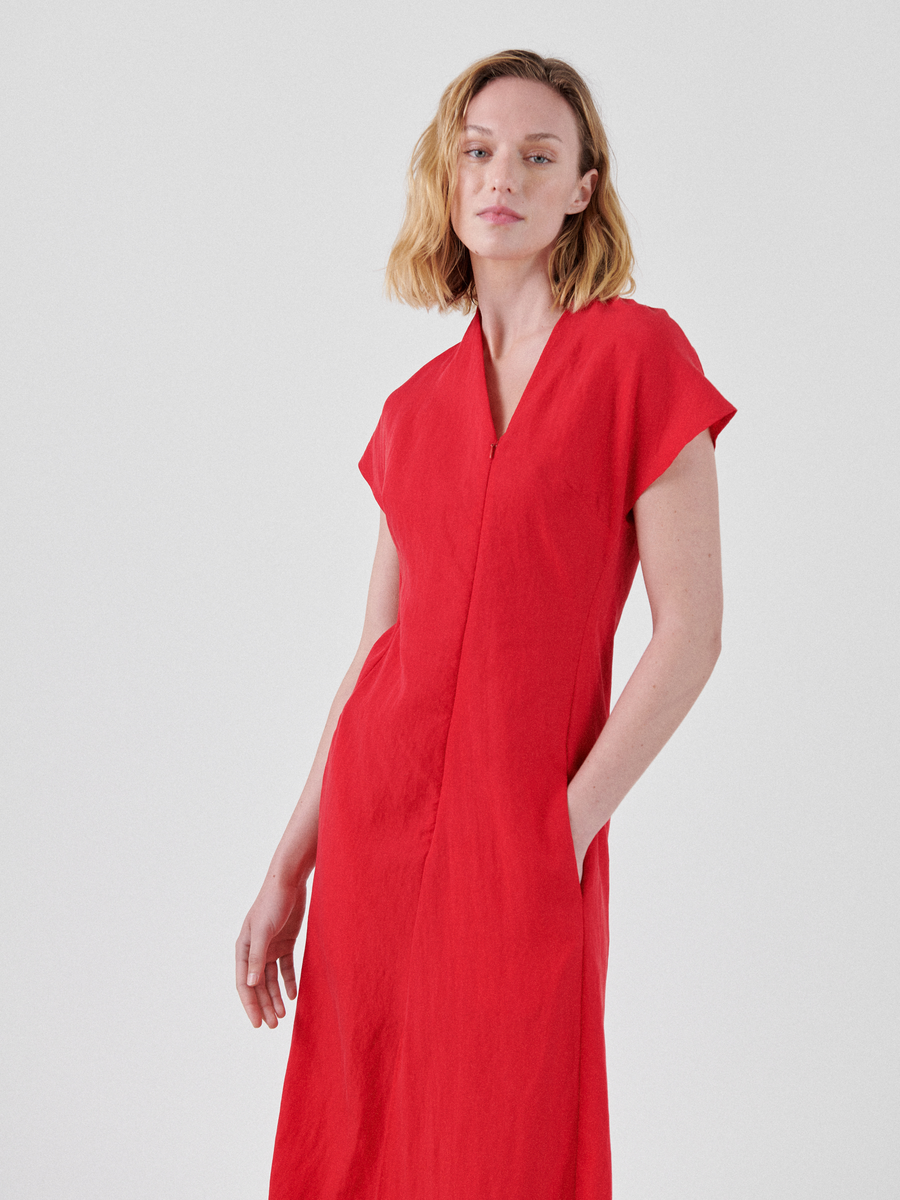 A woman with shoulder-length wavy blonde hair is wearing a bright red, short-sleeved Long Silent Dress by Zero + Maria Cornejo with an asymmetrical V-neckline. She stands against a plain white background with her hands in the pockets of the draped skirt, looking slightly to the side.