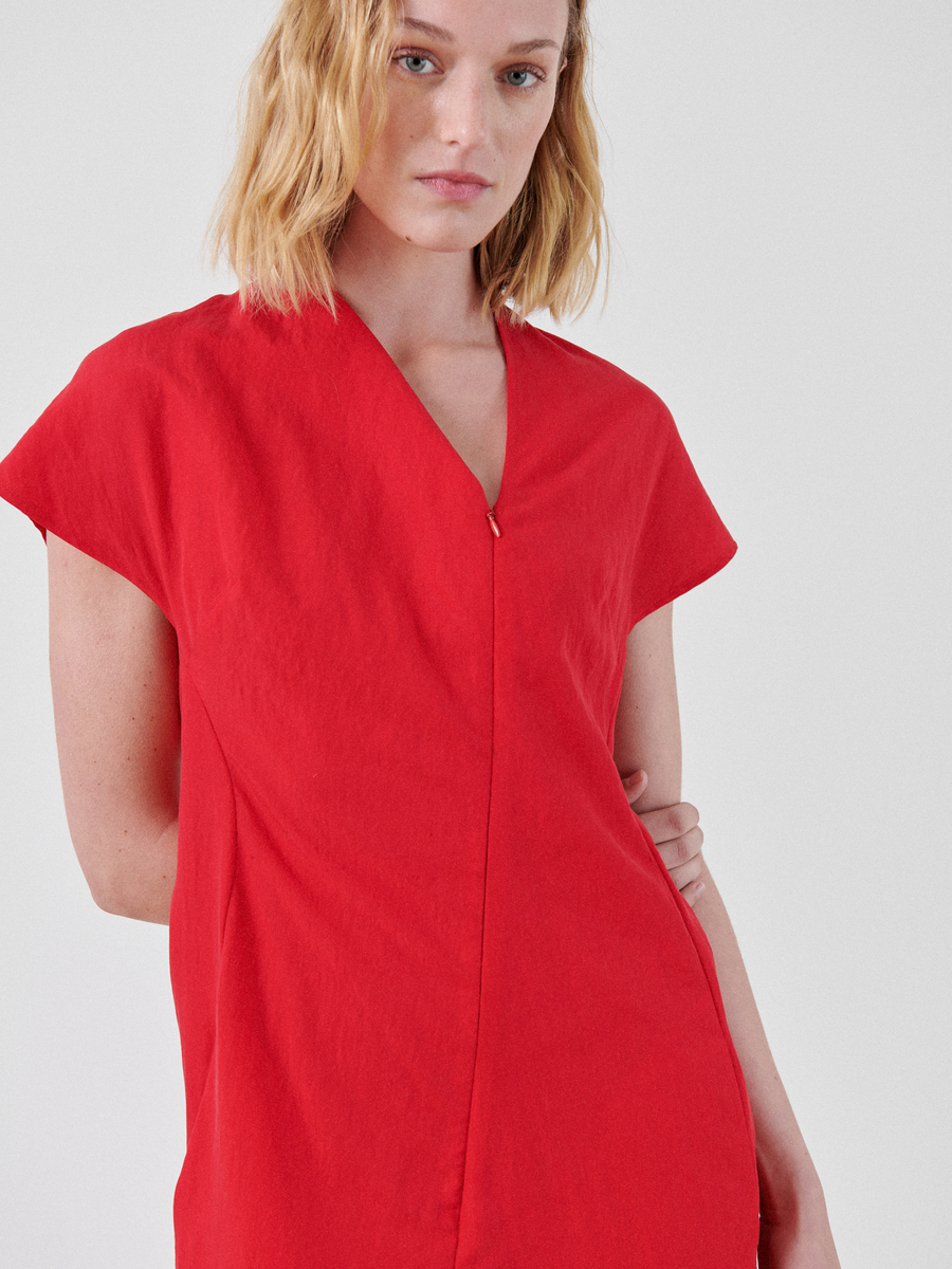 A person with shoulder-length blonde hair is wearing a red Long Silent Dress by Zero + Maria Cornejo. They are standing against a plain white background, looking directly at the camera with a neutral expression.