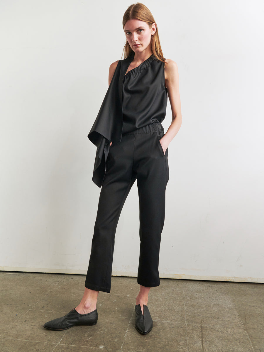 A woman with light skin and long light brown hair stands against a plain white background, wearing a stylish asymmetrical black sleeveless top paired with Slim Elli Pant by Zero + Maria Cornejo. Her relaxed leg pants feature hidden pockets, adding to her outfit's sleek and practical appeal.